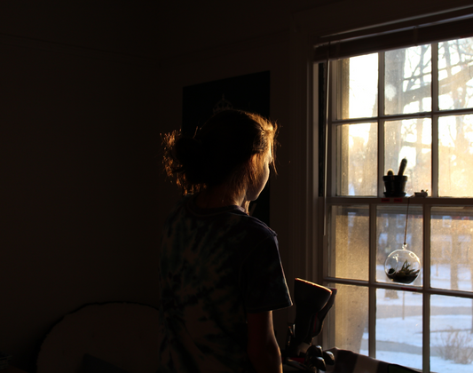 Silhouetted woman standing in a dimly lit room, gazing out a sunlit window, symbolizing isolation, hope, and the emotional struggle of human trafficking survivors.