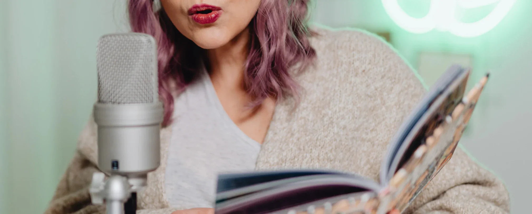 Woman with purple hair reading from an open book into a studio microphone, suggesting storytelling or audiobook narration.