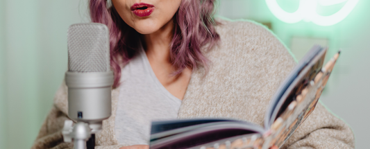 woman with purple hair reading from an open book into a studio microphone, suggesting storytelling or audiobook narration
