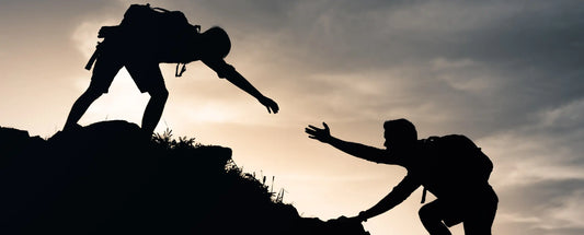 Silhouette of one hiker reaching down to help another climb a steep hillside at sunset, symbolizing support, teamwork, and helping others overcome challenges.