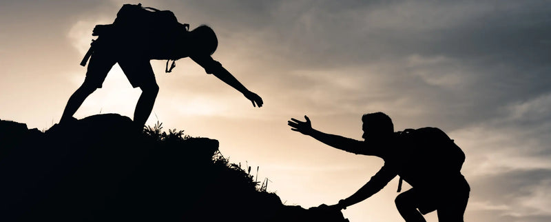 Silhouette of one hiker reaching down to help another climb a steep hillside at sunset, symbolizing support, teamwork, and helping others overcome challenges.