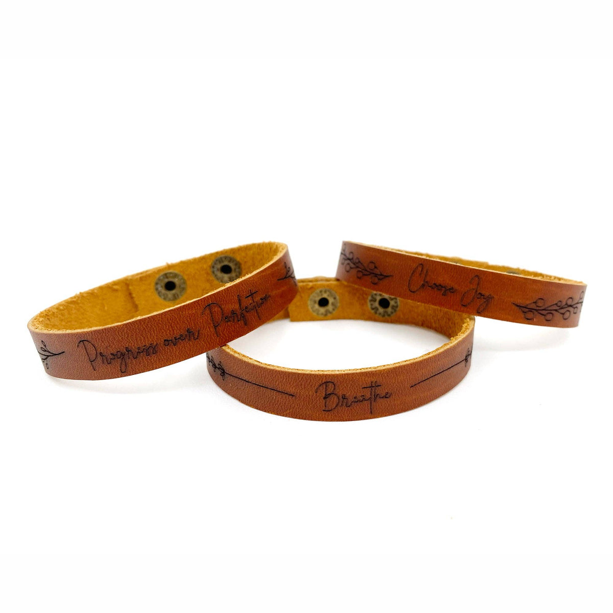 Three tan leather bracelets with black engraved text and decorative line details, displayed on a clean white background.