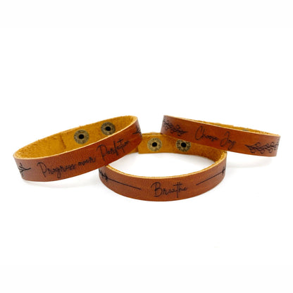 Three tan leather bracelets with black engraved text and decorative line details, displayed on a clean white background.
