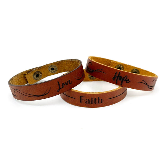 Three brown leather bracelets stacked together on a white background, each engraved with a different word: 'Love', 'Faith', and 'Hope' with black script and decorative flourishes.