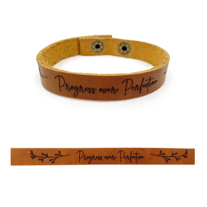 A tan leather bracelet with the engraved phrase 'Progress over Perfection' in black script, shown on a white background with a smaller horizontal close-up inset of the same design.
